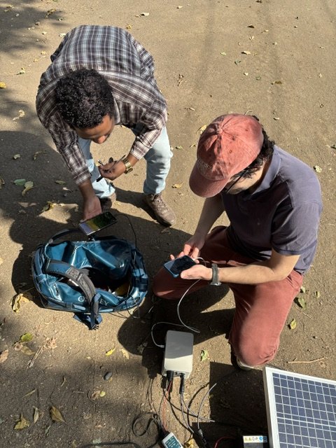 Two people crouched on the ground beside a solar panel, a measurement enclosure and cabling, reading values from their phones