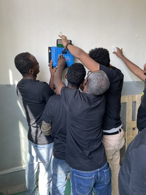 A group of trainees gathered around a solar charge controller being mounted on a wall